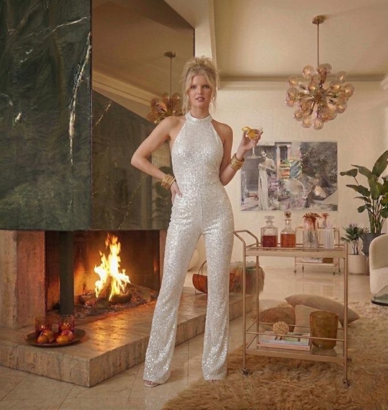 May include: A sleeveless white sequined jumpsuit featuring a high halter neckline and a fitted bodice that transitions into straight-leg trousers. The garment is covered in reflective white sequins that create a shimmering effect across the entire surface.