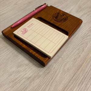 Op de afbeelding: Een houten bureau-organizer met een ingebouwd notitieblok en een roze pen. Op het notitieblok staat "Daily plan". De organizer is gemaakt van donkerbruin hout en heeft een uitgesneden ontwerp.