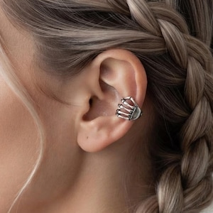 May include: A silver-toned ear cuff shaped like a skeletal hand with five distinct fingers wrapping around the cartilage of the ear. The metal features an oxidized finish in the crevices to highlight the bone structure and anatomical details.