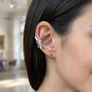 May include: A silver-coloured dragonfly ear cuff worn on the upper cartilage and a small gold-coloured stud earring in the earlobe. The dragonfly design features detailed wing patterns and a slender body, while the stud is a simple, solid geometric shape.