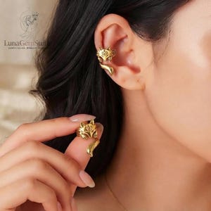May include: A gold-toned ear cuff shaped like a stylized fox with pointed ears and a curved tail. The fox design features intricate engraved details on the face and body. One piece is worn on the upper ear cartilage, while a matching piece is held.