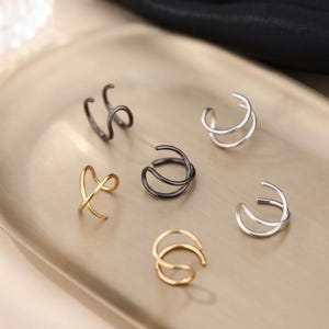 May include: A collection of six ear cuffs in various metallic finishes, including gold, silver, and dark gunmetal. Each piece features a simple, open-loop design with smooth, rounded edges, arranged in a cluster to showcase the different colors.