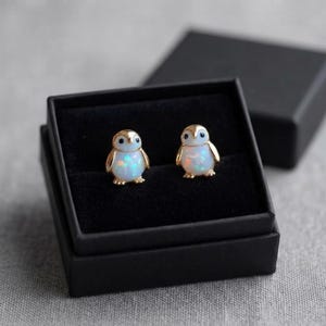 May include: A pair of stud earrings shaped like penguins, featuring gold-toned metal outlines and iridescent white opal-like bodies with small black eyes, presented inside a square black jewelry box with a soft black velvet interior lining.