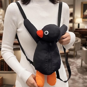 May include: A black backpack shaped like a penguin with a bright orange beak and feet. The penguin features a white eye with a black pupil, a central vertical zip closure on its body, and adjustable black shoulder straps for carrying.