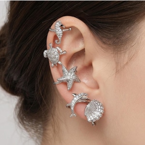 May include: A set of silver-toned ear cuffs and earrings featuring marine life motifs. The collection includes a seahorse, a sea turtle, a starfish, a dolphin, and a seashell, all encrusted with small clear crystals that catch the light.