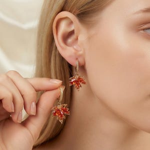 May include: Gold hoop earrings featuring a dangling maple leaf charm. The leaf is encrusted with vibrant orange and red crystals, while the hoop is adorned with small clear stones. The design captures the essence of autumn with its warm colors.