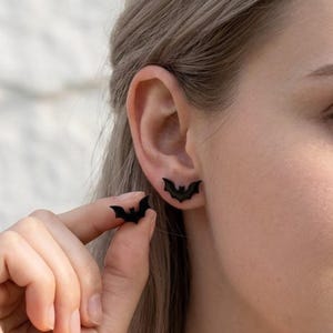 Peut inclure: Une paire de boucles d'oreilles à clous noir mat en forme de petites chauves-souris aux ailes déployées. Une boucle d'oreille est placée sur le lobe de l'oreille, tandis que l'autre est tenue entre le pouce et l'index pour montrer sa forme.