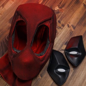 K&ouml;nnte beinhalten: Eine strukturierte rote Stoffmaske mit zwei gro&szlig;en Augen&ouml;ffnungen und einer roten Stoff-Halsabdeckung, begleitet von zwei separaten schwarzen Augenplatten-Eins&auml;tzen mit wei&szlig;en ovalen Linsen, entworfen als Zubeh&ouml;r f&uuml;r ein Ganzkopfkost&uuml;m.