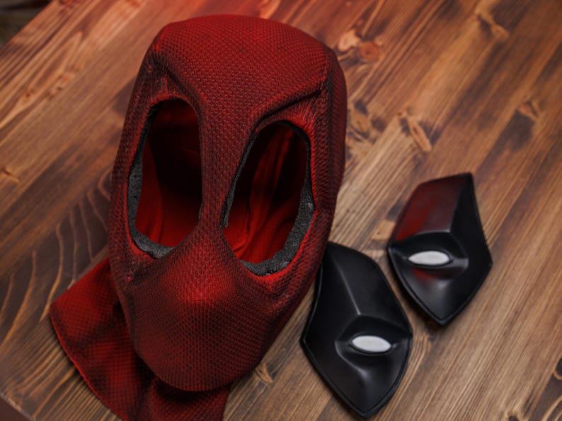 K&ouml;nnte beinhalten: Eine strukturierte rote Stoffmaske mit zwei gro&szlig;en Augen&ouml;ffnungen und einer roten Stoff-Halsabdeckung, begleitet von zwei separaten schwarzen Augenplatten-Eins&auml;tzen mit wei&szlig;en ovalen Linsen, entworfen als Zubeh&ouml;r f&uuml;r ein Ganzkopfkost&uuml;m.