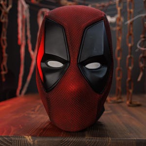 K&ouml;nnte beinhalten: Eine rot-schwarze Vollkopfmaske mit einer strukturierten Stoffoberfl&auml;che, glatten schwarzen Augenpartien mit wei&szlig;en Netzgl&auml;sern und einem markanten kantigen Design. Die Maske zeigt eine leuchtend rote Farbe auf dem Hauptteil mit schwarzen Akzenten.