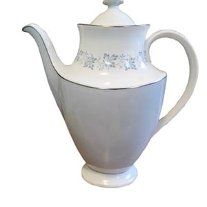 May include: A white porcelain coffee pot featuring a tapered body, a curved handle, and a spout. The lid has a pointed finial. A band of light blue maple leaf patterns encircles the upper section, and silver trim accents the rim and lid.