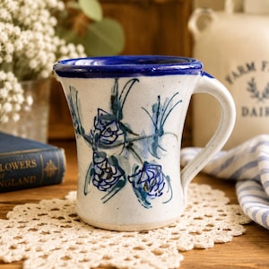 May include: A ceramic mug with a speckled off-white base and a vibrant blue rim. The exterior features hand-painted blue and green floral motifs. It has a curved handle on the right side and a slightly flared top edge, showcasing a classic pottery design.