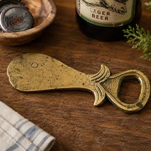 May include: A brass bottle opener shaped like a whale with a textured finish. The whale-shaped tool features a simple circular eye and a tail section that functions as the opening mechanism for removing metal bottle caps from glass beverage bottles.