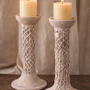 May include: Two off-white candle holders with lit candles. The holders have a textured, woven design on the stems and a wide base. The candles are ivory-colored and cylindrical. The image includes a "Made in PORTUGAL" label.