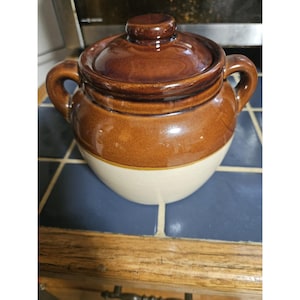 May include: A two-handled ceramic bean pot with a glossy brown glazed lid and upper body, transitioning to a matte cream-colored base. The pot features a rounded shape with two side handles and a small knob on the center of the lid.