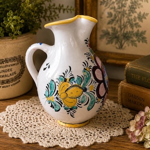 May include: A white ceramic pitcher featuring a hand-painted floral design in yellow, teal, and dark purple. The pitcher has a curved handle, a flared spout with a yellow rim, and a rounded body. The painted motifs include stylized petals and leaves.