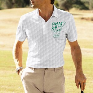 K&ouml;nnte beinhalten: Wei&szlig;es Poloshirt mit Golfballmuster und einer gr&uuml;nen Grafik von Mickey Mouse beim Golfspielen, mit dem Text "MM 1928". Das Shirt hat einen Kragen und kurze &Auml;rmel. Die Person tr&auml;gt eine khakifarbene Hose und eine Uhr.