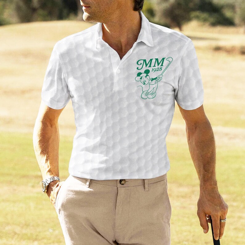 K&ouml;nnte beinhalten: Wei&szlig;es Poloshirt mit Golfballmuster und einer gr&uuml;nen Grafik von Mickey Mouse beim Golfspielen, mit dem Text "MM 1928". Das Shirt hat einen Kragen und kurze &Auml;rmel. Die Person tr&auml;gt eine khakifarbene Hose und eine Uhr.