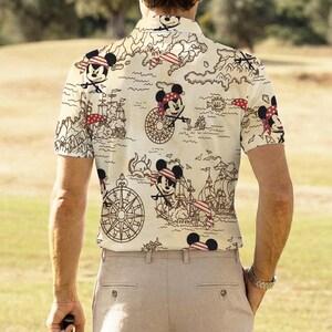 May include: Short-sleeved collared shirt with a cream background featuring a pirate-themed design. The design includes cartoon images of Mickey Mouse wearing a pirate bandana, pirate ships, compasses, and a treasure map.