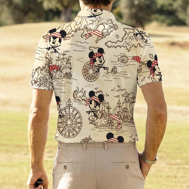 May include: Short-sleeved collared shirt with a cream background featuring a pirate-themed design. The design includes cartoon images of Mickey Mouse wearing a pirate bandana, pirate ships, compasses, and a treasure map.