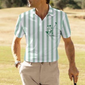 May include: A polo shirt with vertical stripes in white and light green. The shirt features a graphic of a cartoon mouse playing golf, with the name "Smiths" below. The person is wearing khaki pants and a watch.