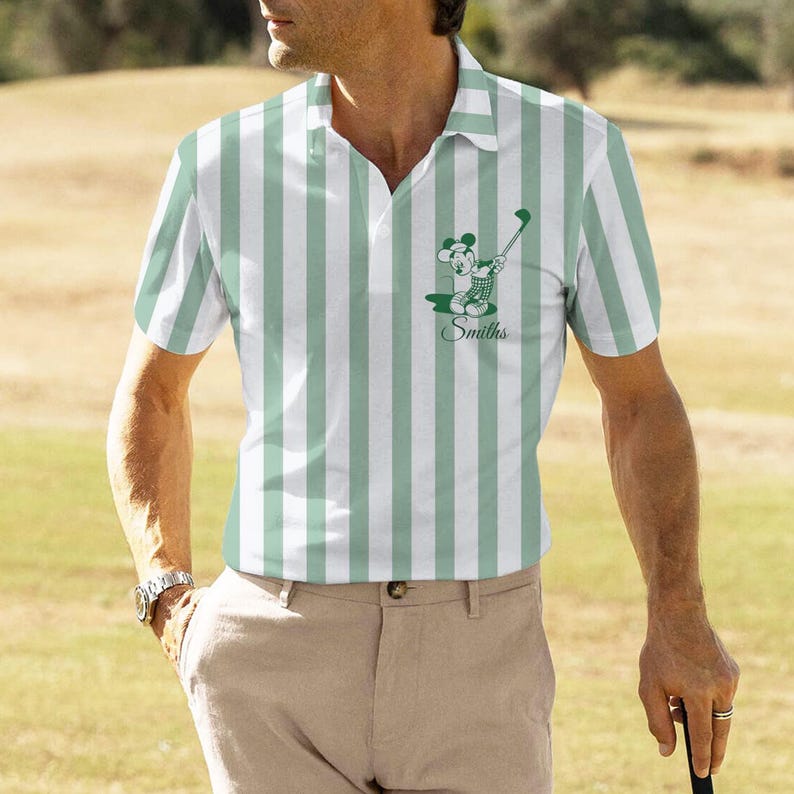 May include: A polo shirt with vertical stripes in white and light green. The shirt features a graphic of a cartoon mouse playing golf, with the name "Smiths" below. The person is wearing khaki pants and a watch.