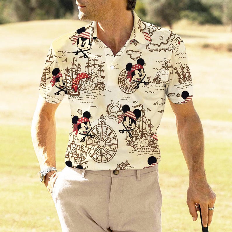 May include: Cream-colored polo shirt featuring a repeating pattern of Mickey Mouse as a pirate, ships, and nautical maps. The shirt has a collared neckline and short sleeves. The design incorporates red, black, and brown elements.