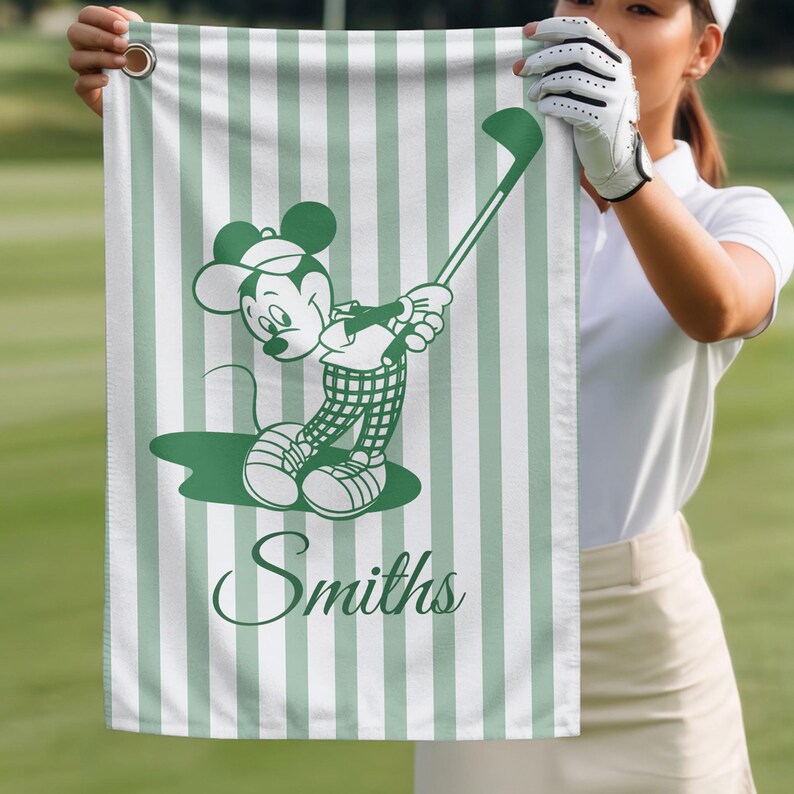 May include: A golf towel with a green and white striped pattern. The towel features a green illustration of Mickey Mouse swinging a golf club. The name "Smiths" is printed in a script font at the bottom of the towel.