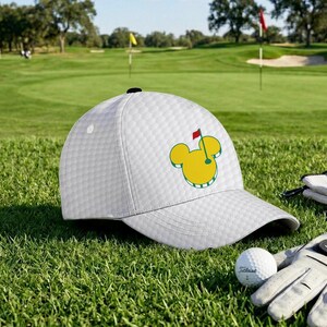 May include: White golf cap with a textured pattern and a yellow and green graphic of a golf flag and Mickey Mouse ears. A golf ball and glove are visible on the green grass.
