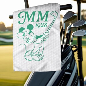 K&ouml;nnte beinhalten: Wei&szlig;es Golfhandtuch mit gr&uuml;ner Umrisszeichnung von Mickey Mouse beim Golfspielen und dem Text "MM 1928". Das Handtuch h&auml;ngt an einem Golfwagen, im Hintergrund Golfschl&auml;ger.
