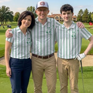 May include: Three people wearing matching green and white striped polo shirts with a Mickey Mouse graphic and the name "Smiths" on the chest. One person is wearing a baseball cap. They are standing on a golf course.