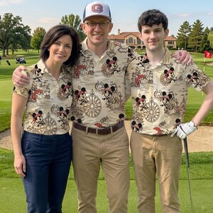 May include: Three people wearing matching beige polo shirts with a nautical Mickey Mouse print, posing on a golf course. The shirts feature a repeating pattern of Mickey Mouse as a pirate, ships, and compasses. One person wears a baseball cap.