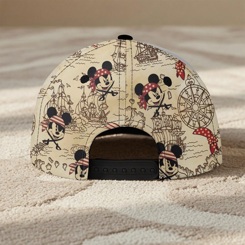 May include: A beige baseball cap featuring a pirate-themed design with cartoon Mickey Mouse characters, ships, and a compass. The cap has a black brim and a snapback closure.