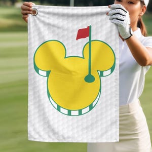 May include: White golf towel featuring a yellow Mickey Mouse head design with a green outline. The design includes a golf flag with a red flag and a green pole. The towel has a honeycomb pattern.