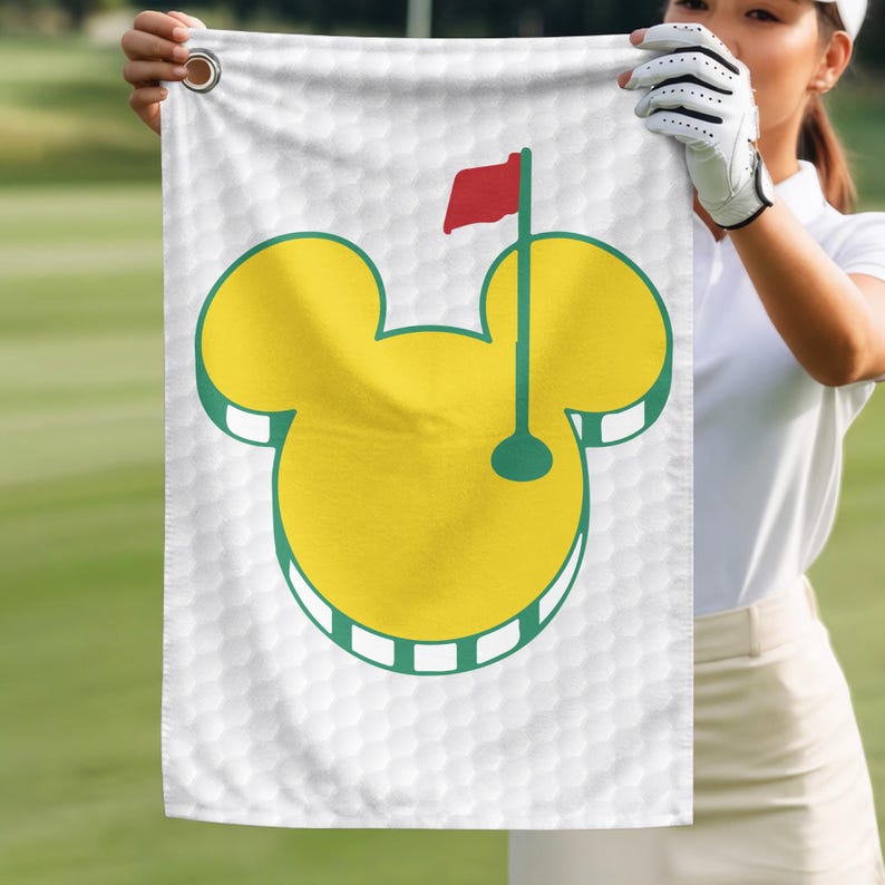 May include: White golf towel featuring a yellow Mickey Mouse head design with a green outline. The design includes a golf flag with a red flag and a green pole. The towel has a honeycomb pattern.