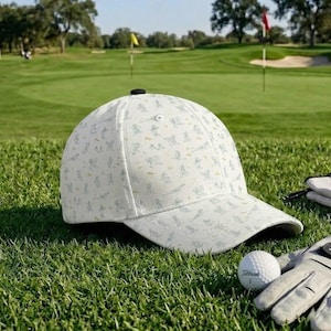 Może przedstawiać: Biała czapka bejsbolowa z motywem golfowym, czarnym guzikiem na g&oacute;rze i zakrzywionym daszkiem. Piłka golfowa i rękawica leżą na trawie. Tło przedstawia pole golfowe z zieloną trawą.