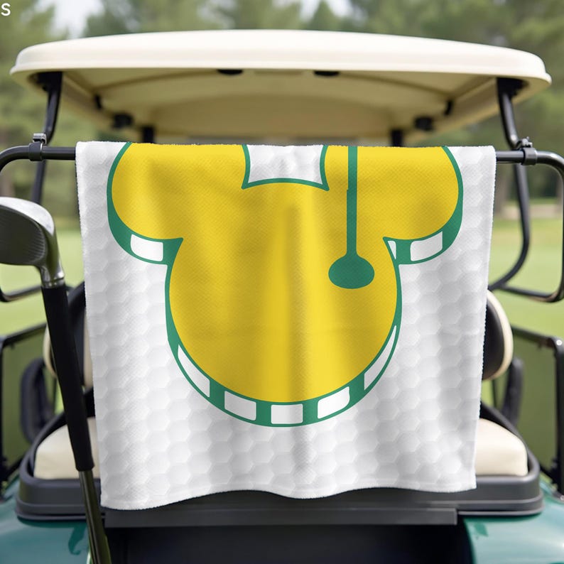 May include: White golf towel with a yellow and green golf-themed design hanging on a golf cart. The towel features a yellow golf ball shape with green accents and a golf club design. The background has a golf ball pattern.