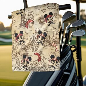 May include: A beige golf towel featuring a pirate-themed map design with cartoon Mickey Mouse characters. The map includes ships, compasses, and red accents. The towel is hanging on a golf cart.