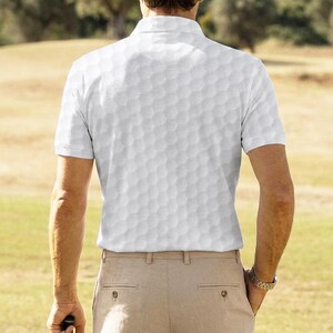 K&ouml;nnte beinhalten: Wei&szlig;es Poloshirt mit Golfballmuster. Das Shirt hat kurze &Auml;rmel und einen Kragen. Die Person tr&auml;gt eine beige Hose und eine Uhr. Der Hintergrund ist ein Golfplatz.