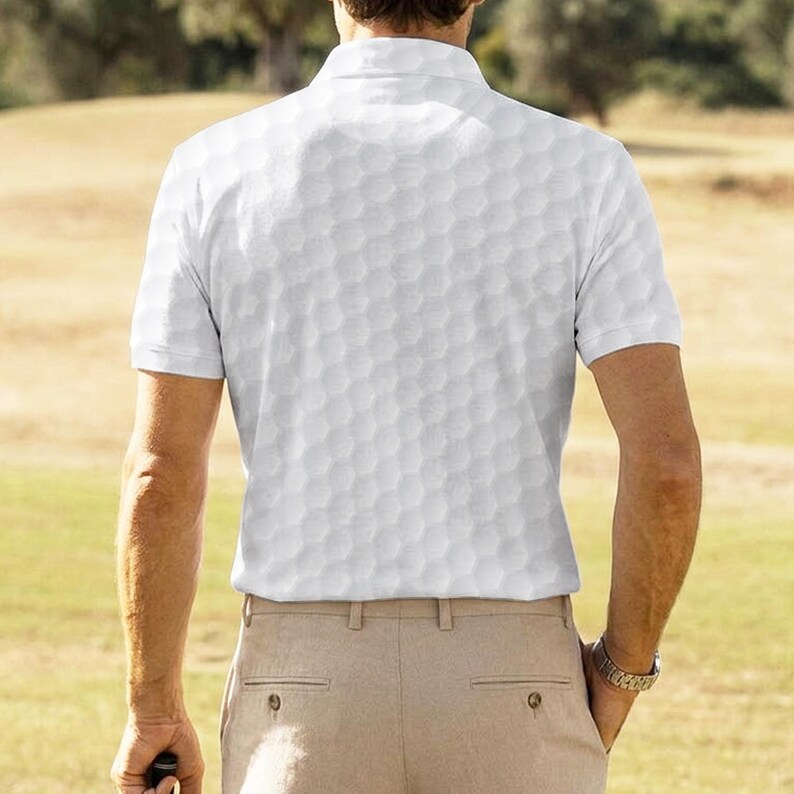 K&ouml;nnte beinhalten: Wei&szlig;es Poloshirt mit Golfballmuster. Das Shirt hat kurze &Auml;rmel und einen Kragen. Die Person tr&auml;gt eine beige Hose und eine Uhr. Der Hintergrund ist ein Golfplatz.