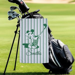 May include: A green and white striped golf towel featuring a cartoon character in a golf pose. The character is wearing a golf outfit and holding a golf club. The name "Smiths" is printed in a script font on the towel. The towel is hanging on a golf bag.