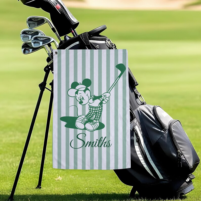 May include: A green and white striped golf towel featuring a cartoon character in a golf pose. The character is wearing a golf outfit and holding a golf club. The name "Smiths" is printed in a script font on the towel. The towel is hanging on a golf bag.