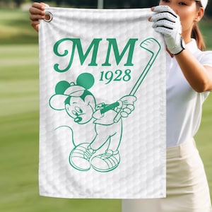 K&ouml;nnte beinhalten: Wei&szlig;es Golfhandtuch mit gr&uuml;ner Illustration einer Cartoon-Maus, die einen Golfschl&auml;ger schwingt. Das Handtuch hat die Initialen "MM" und das Jahr "1928" in Gr&uuml;n aufgedruckt. Der Hintergrund hat ein Golfballmuster.