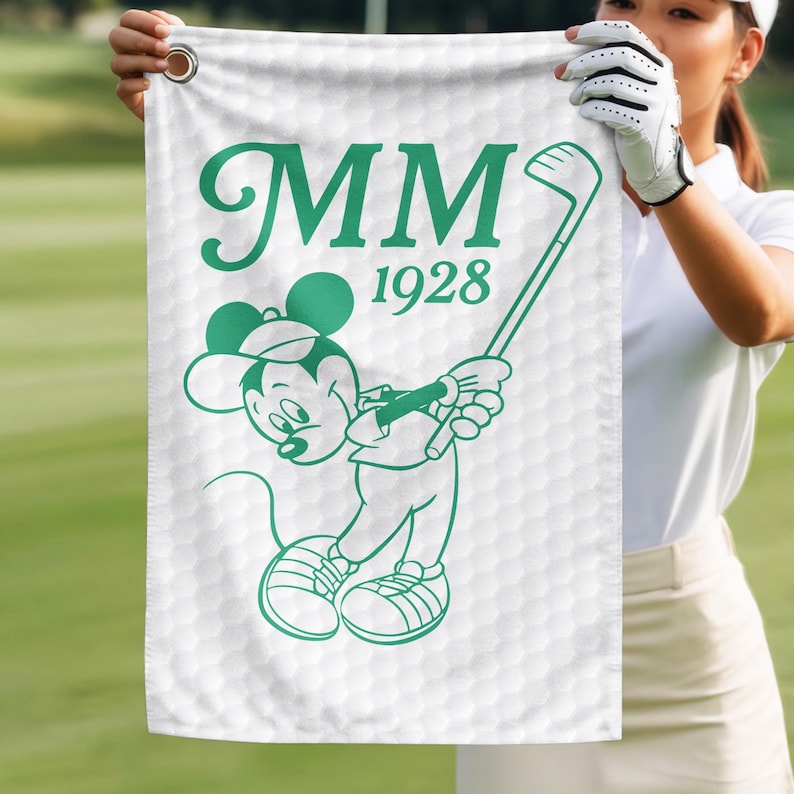K&ouml;nnte beinhalten: Wei&szlig;es Golfhandtuch mit gr&uuml;ner Illustration einer Cartoon-Maus, die einen Golfschl&auml;ger schwingt. Das Handtuch hat die Initialen "MM" und das Jahr "1928" in Gr&uuml;n aufgedruckt. Der Hintergrund hat ein Golfballmuster.