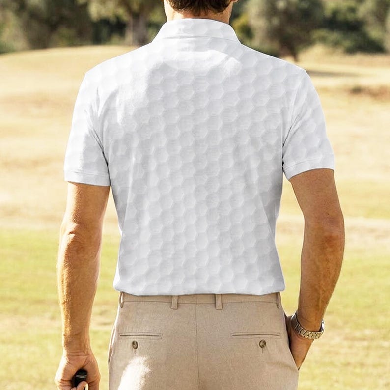 May include: White polo shirt with a golf ball pattern, paired with khaki pants. The shirt has short sleeves and a collared neckline. A watch is visible on the wrist. The person is standing on a grassy area.