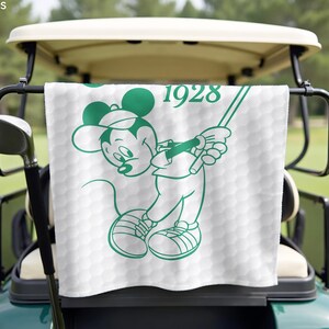 K&ouml;nnte beinhalten: Wei&szlig;es Golfhandtuch mit gr&uuml;ner Umrisszeichnung von Mickey Mouse beim Golfspielen. Das Handtuch hat ein Wabenmuster und die Jahreszahl "1928" in Gr&uuml;n. Es h&auml;ngt an einem Golfwagen.