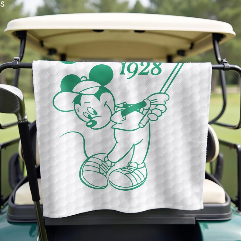 K&ouml;nnte beinhalten: Wei&szlig;es Golfhandtuch mit gr&uuml;ner Umrisszeichnung von Mickey Mouse beim Golfspielen. Das Handtuch hat ein Wabenmuster und die Jahreszahl "1928" in Gr&uuml;n. Es h&auml;ngt an einem Golfwagen.