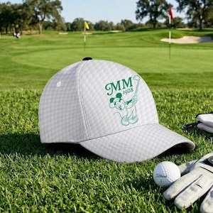 K&ouml;nnte beinhalten: Wei&szlig;e Golfkappe mit strukturiertem Muster, mit einer gr&uuml;nen Grafik von Mickey Mouse beim Golfschl&auml;ger schwingen, mit den Initialen "MM" und dem Jahr "1928". Ein Golfball und ein Handschuh sind auf dem gr&uuml;nen Gras zu sehen.