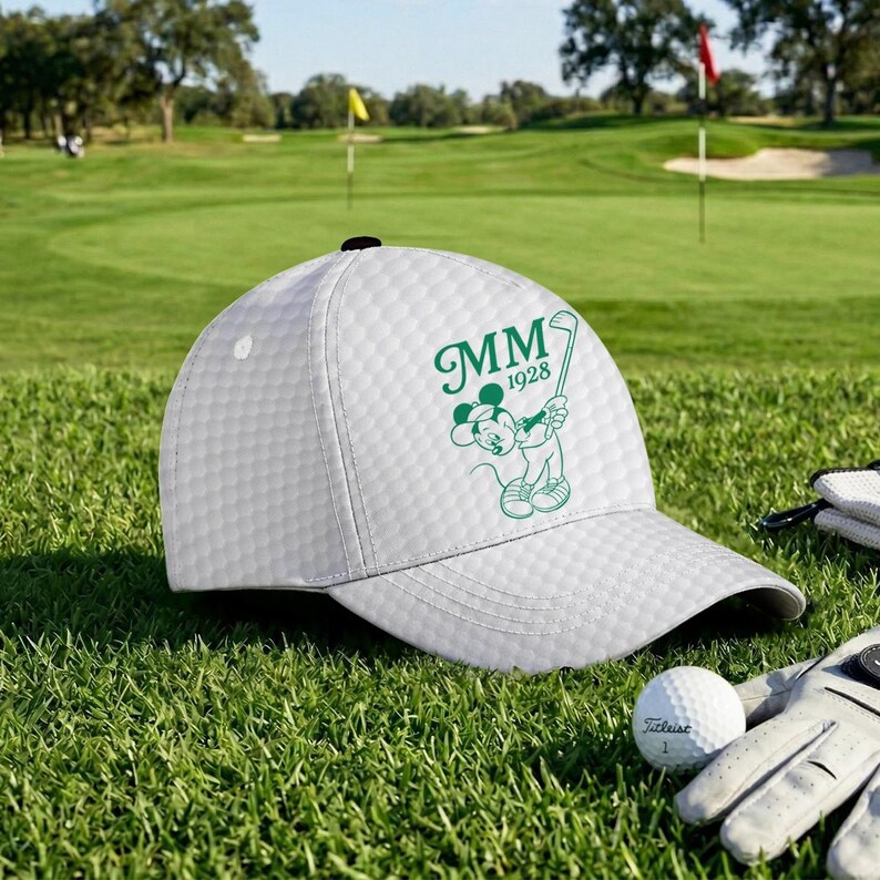 K&ouml;nnte beinhalten: Wei&szlig;e Golfkappe mit strukturiertem Muster, mit einer gr&uuml;nen Grafik von Mickey Mouse beim Golfschl&auml;ger schwingen, mit den Initialen "MM" und dem Jahr "1928". Ein Golfball und ein Handschuh sind auf dem gr&uuml;nen Gras zu sehen.