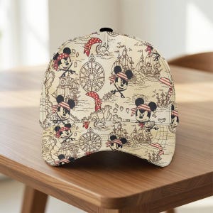 May include: A beige baseball cap featuring a pirate-themed design with cartoon characters, ships, compasses, and red accents. The cap is set on a wooden surface.